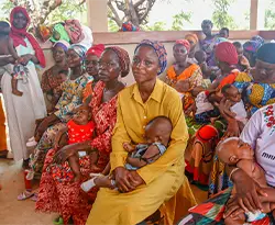 A large grouop of women holding their babies. They're sitting in a waiting area to see a health professional.