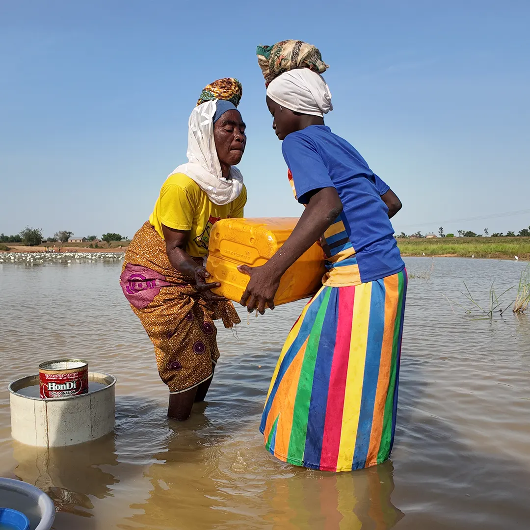 Two women standing in ankle deep water, holding a yellow jerry can.