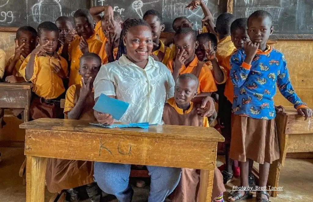 A woman sitting behind a desk surrounded by smiling children in a classroom